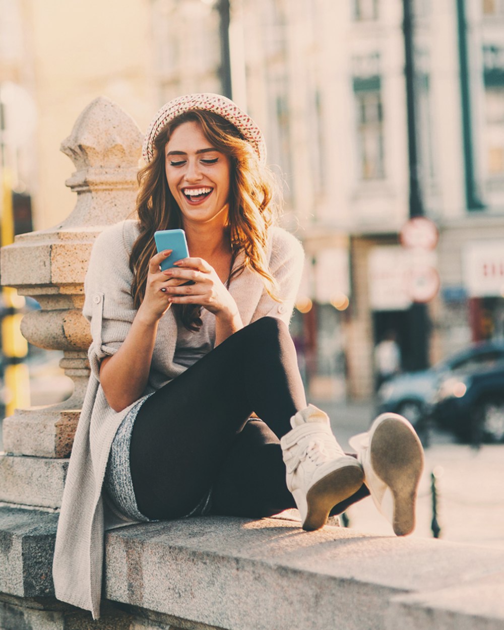 A woman sitting on a stone wall in a city at golden hour, laughing at her phone